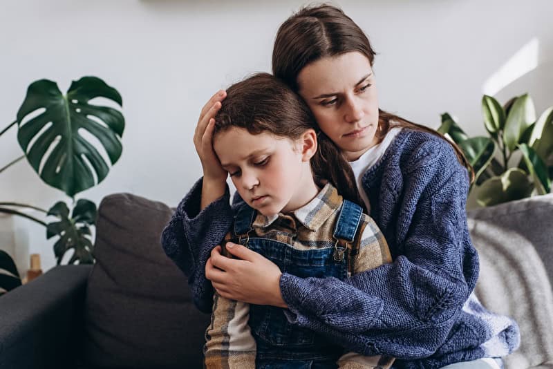 Fürsorgliche junge Mutter umarmt, beruhigt aufgebrachte kleine Tochter, sitzt auf der Couch im Wohnzimmer zu Hause, liebevolle Mama tröstet beleidigtes trauriges Mädchen Kind, drückt Liebe und Unterstützung aus. Konzept der Kinderpsychologen